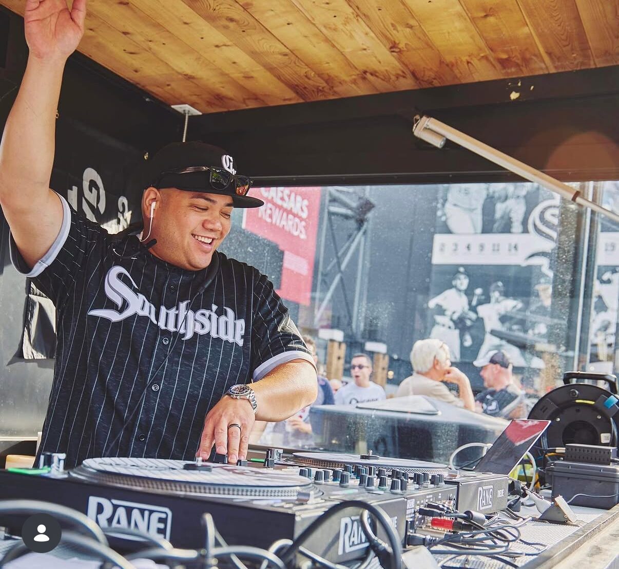A DJ wearing a "Southside" jersey and headphones plays music on a mixer at an outdoor event, raising one hand and smiling.