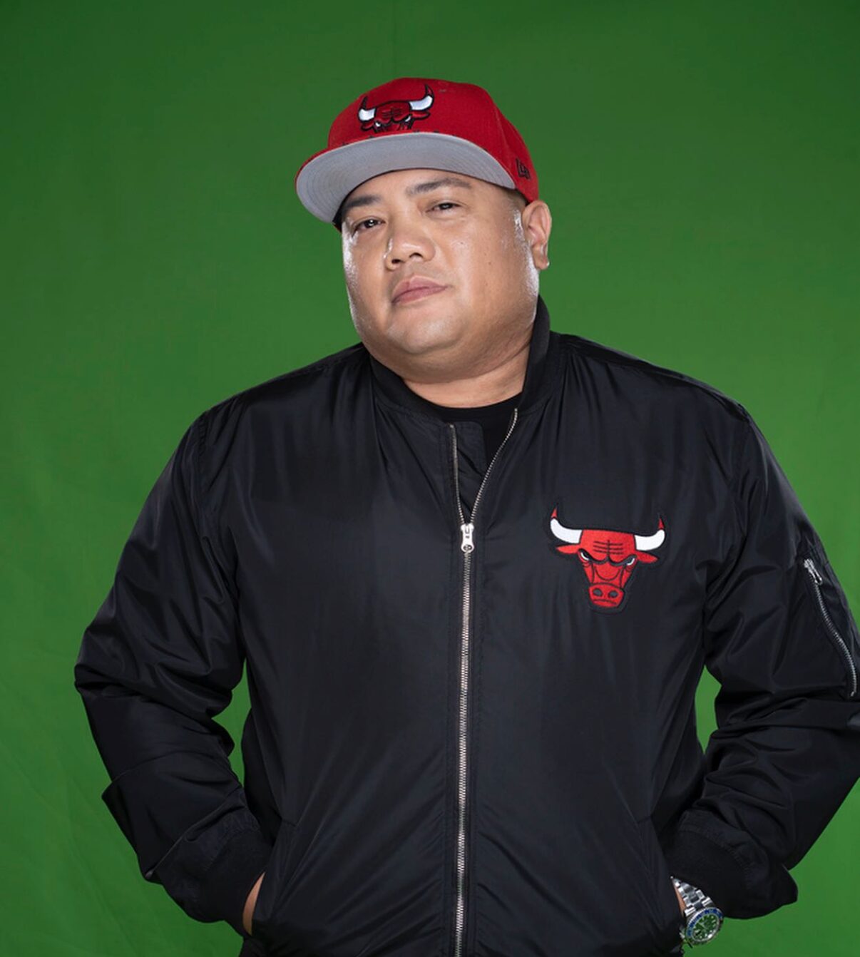 A man stands in front of a green background wearing a black jacket and red cap, both featuring the Chicago Bulls logo.
