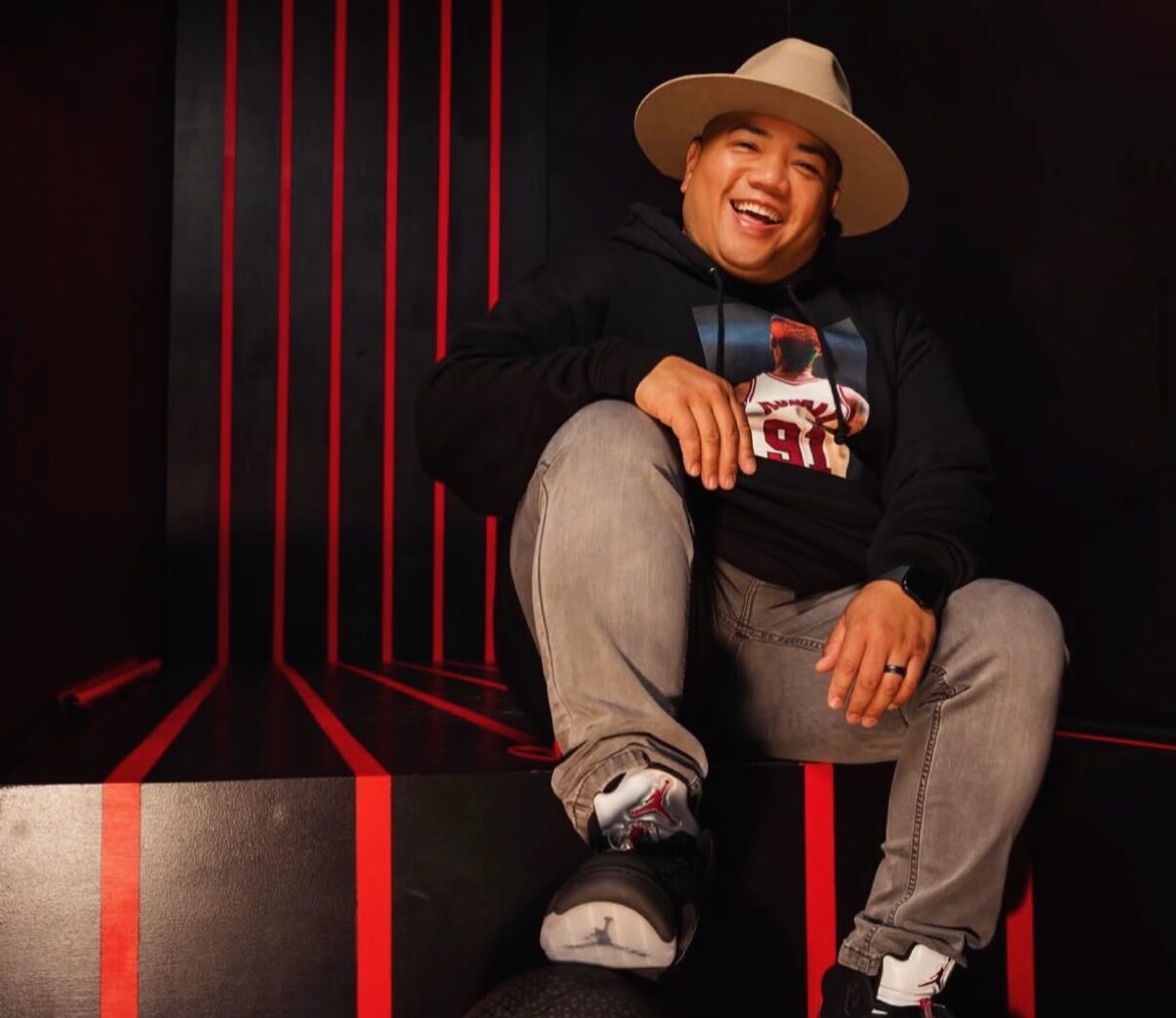 A person wearing a wide-brim hat, hoodie, jeans, and sneakers sits on a black bench with red stripes, smiling at the camera.