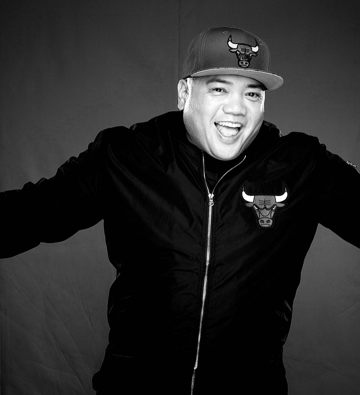 A person wearing a Chicago Bulls cap and jacket smiles with arms outstretched against a plain background. The image is in black and white.