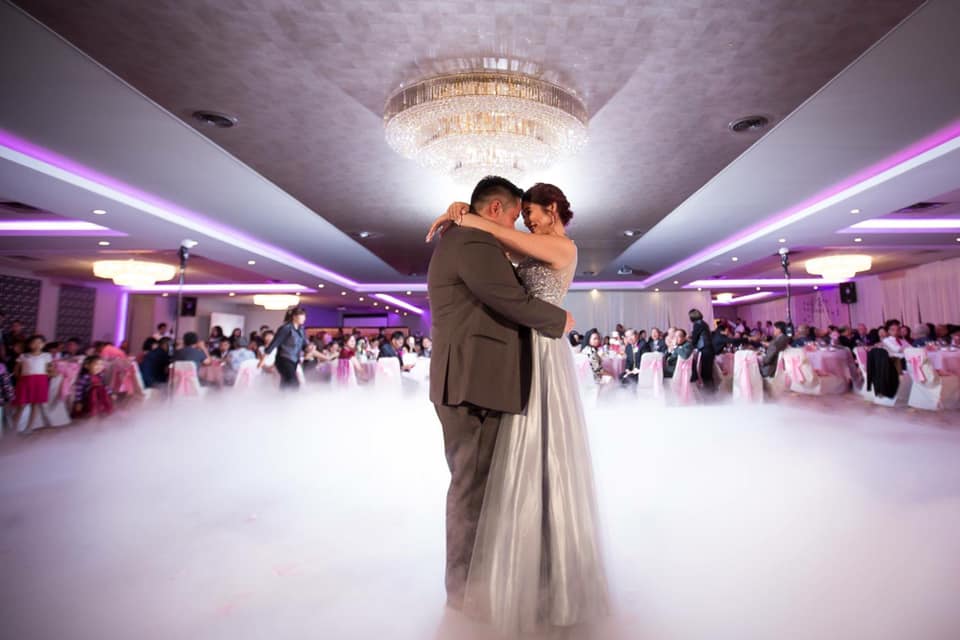 A couple in formal attire shares a dance in the center of a fog-covered ballroom, surrounded by seated guests at a decorated event as the wedding DJ sets the perfect mood.