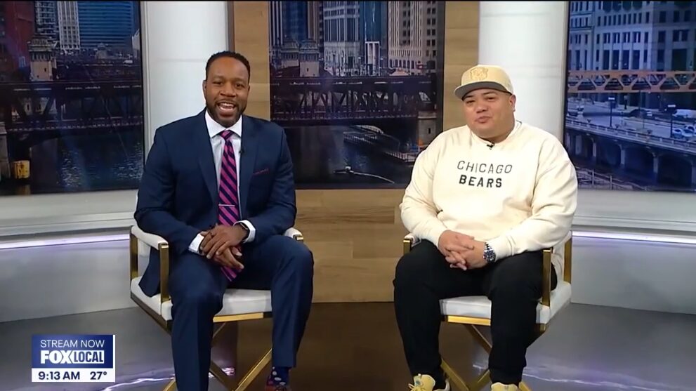 Two men sit in chairs on a TV studio set; one wears a suit and tie, the other sports a "Chicago Bears" sweatshirt and cap. A cityscape glows in the background, capturing the authentic style of momentz productions.