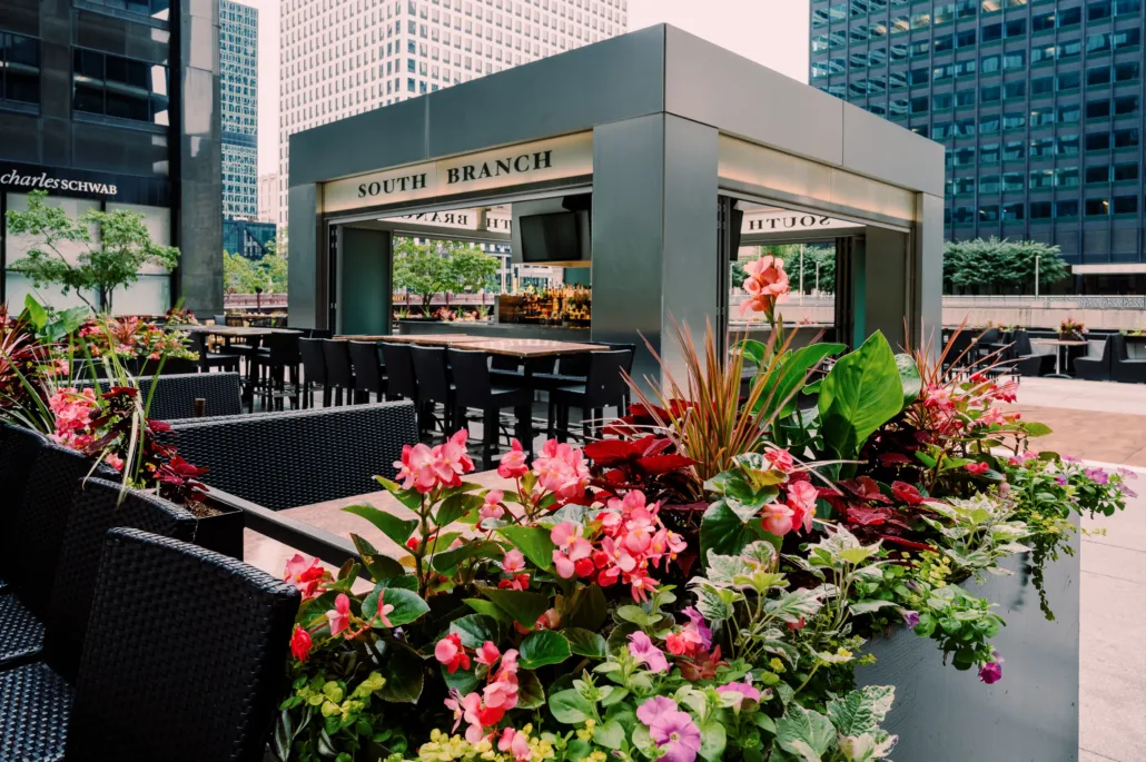 Outdoor dining area with modern seating and colorful flower planters, surrounded by tall office buildings; a bar labeled "South Branch" is at the center, making it a standout among Chicago wedding venues.