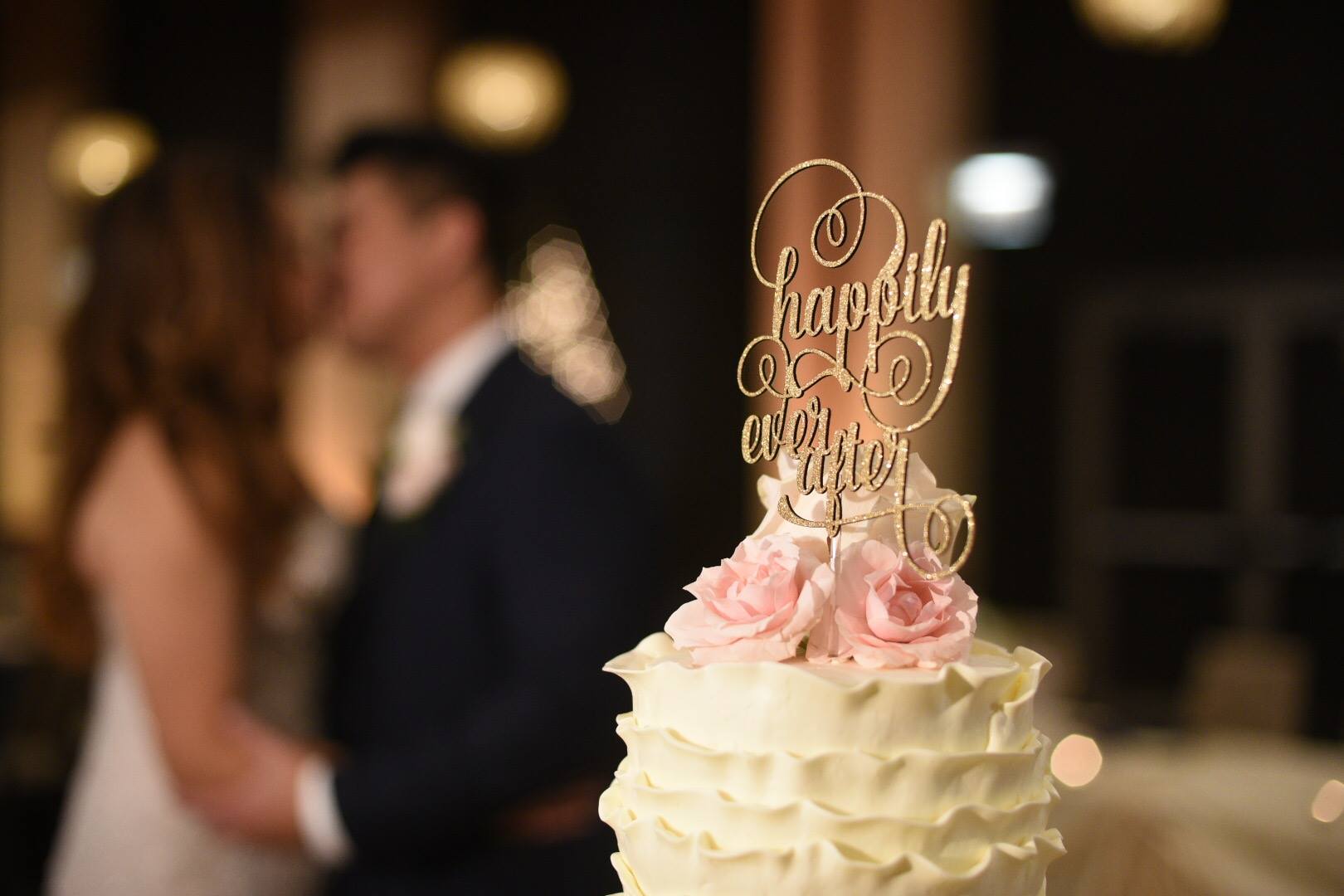 A white wedding cake with pink roses and a "happily ever after" topper in focus; a bride and groom, enjoying the music from their wedding DJ, are blurred in the background.