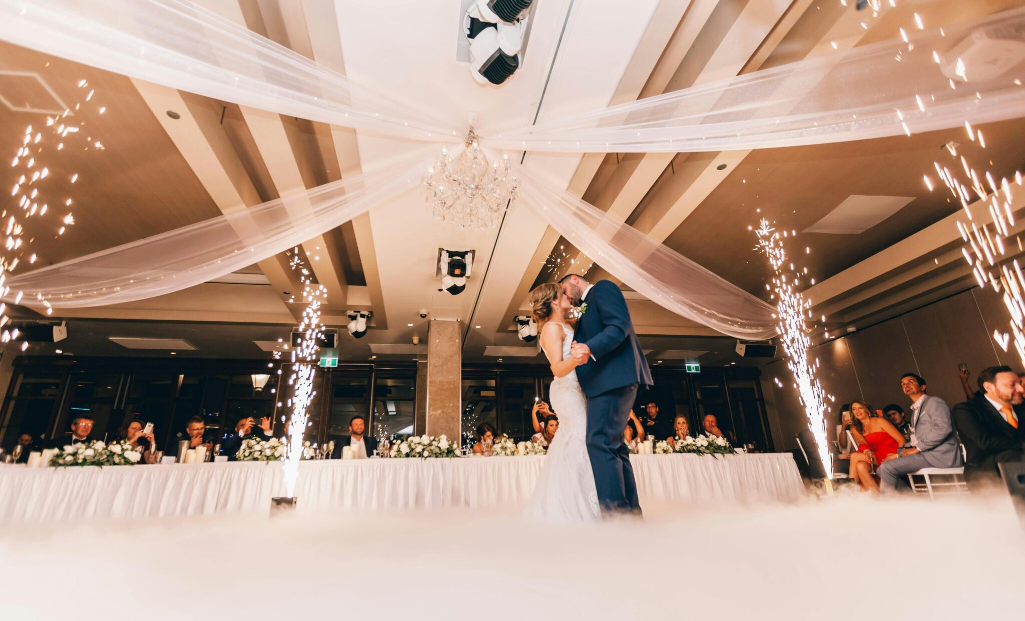 A bride and groom share a first dance under draped fabric and chandeliers, surrounded by guests and indoor fireworks at a decorated wedding reception—don’t miss 5 important tips your wedding DJ wants you to know for moments like these.