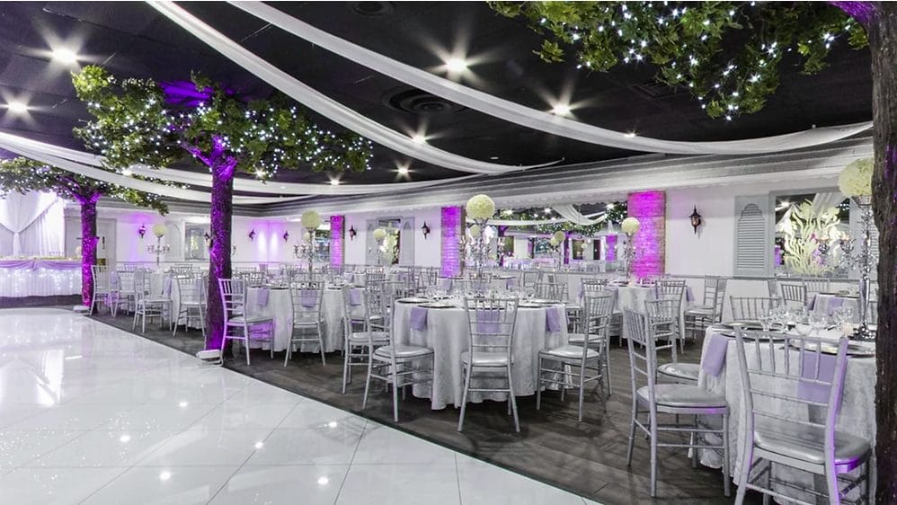Elegant banquet hall decorated with artificial trees and white drapery, featuring round tables with white tablecloths and silver chairs, lit with soft purple lighting.