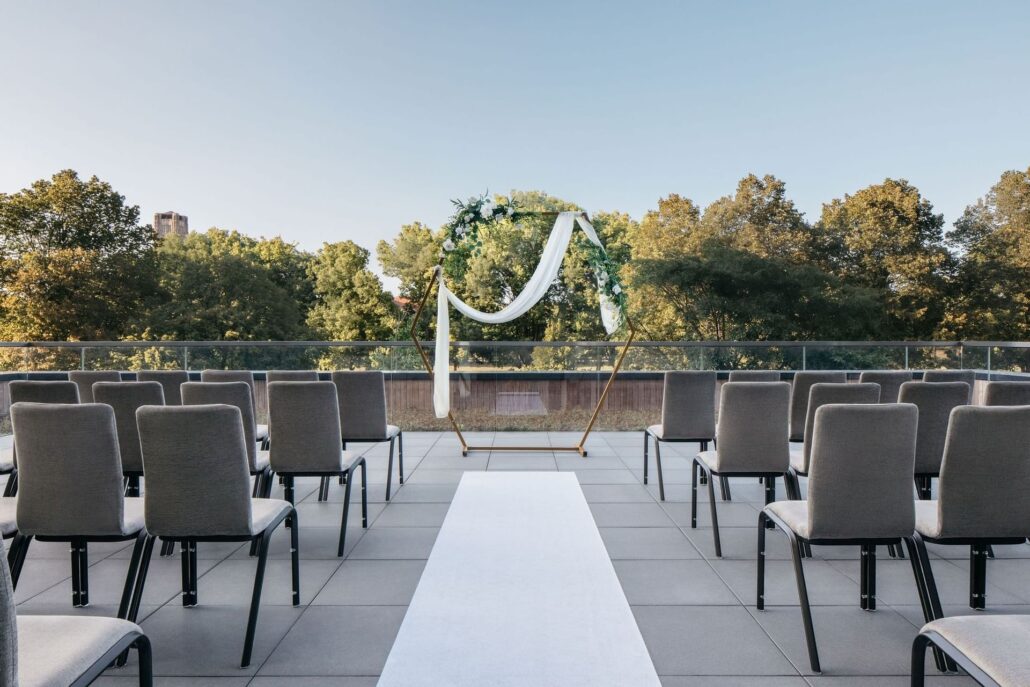 Rows of gray chairs face a geometric wedding arch adorned with greenery and white fabric on an outdoor terrace, offering a modern touch to Chicago wedding venues with scenic views of trees in the background.